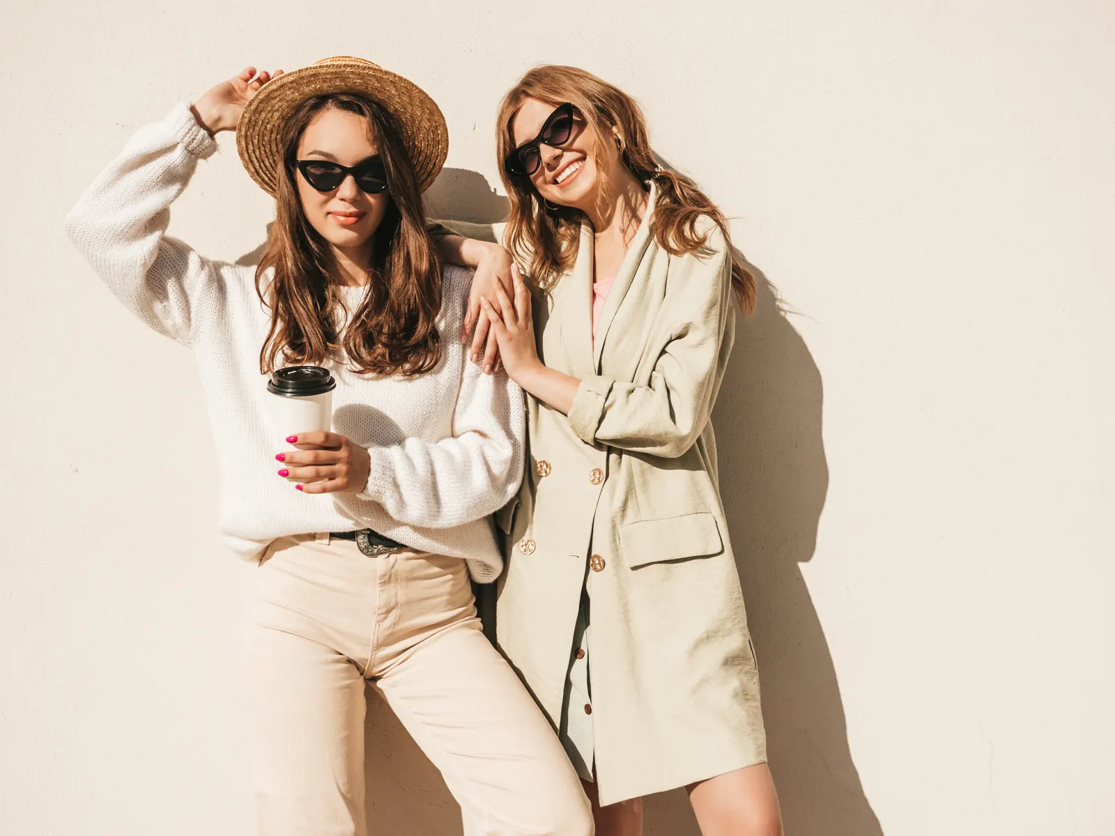 Dos amigas con suéter de punto blanco y blazer crudo, con lentes de sol, apoyadas en una pared clara al sol
