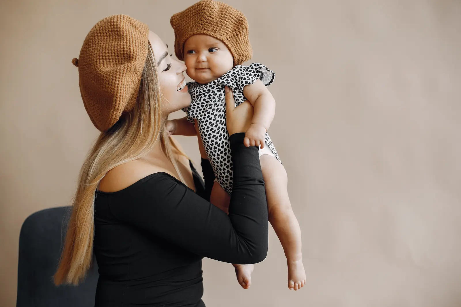 Mãe de blusa preta e boina de tricô caramelo beija o bebê de body estampado e boina igual, em estúdio de fundo bege