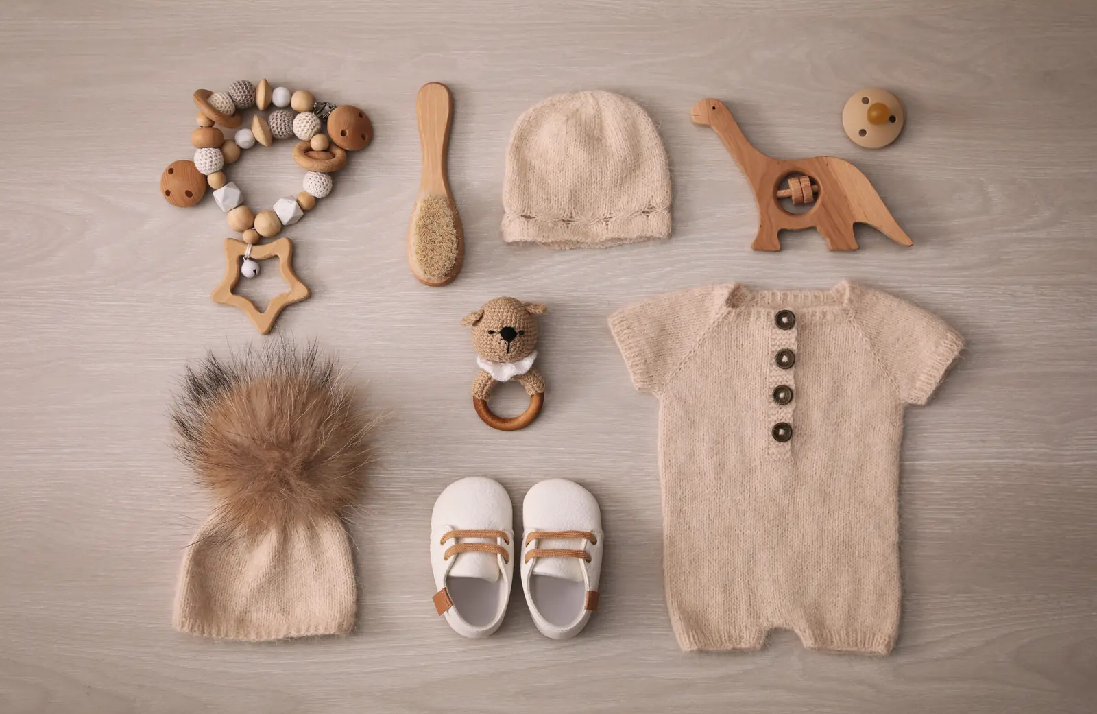 Macaquinho de tricô bege, gorro de tricô, gorro com pompom, tênis branco e mordedores de madeira sobre uma mesa clara